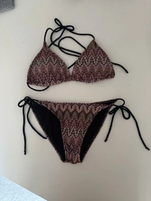 Mönstrad bikini med snörning - Snygg bikini med zigzag-mönster i rosa, svart och beige. Överdelen har trekantskupor och knytband i nacken och ryggen. Underdelen har knyt i sidorna för justerbar passform. Perfekt för stranden eller poolen!