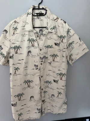 Beige kortärmad skjorta med palmmotiv - Snygg beige skjorta med korta ärmar och tropiskt palmmotiv. Skjortan har knappar framtill och en bröstficka. Perfekt för en avslappnad och somrig stil.