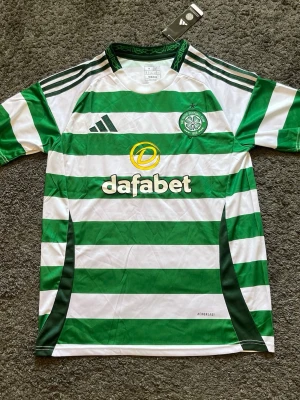 Celtic FC matchtröja  - Säljer en grönvit randig matchtröja från Celtic FC, tillverkad av Adidas. Tröjan har klubbmärke på bröstet, Adidas-logga och sponsortryck på framsidan. Tillverkad i lätt och ventilerande material med slim fit passform. Perfekt för fotbollsträning eller match.