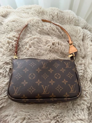  Monogram Pochette - Snygg handväska i klassiskt brunt monogrammönster med guldiga detaljer och läderrem. Väskan har dragkedja upptill och är tillverkad i canvas med läderdetaljer. Perfekt för dig som gillar lyxiga accessoarer och tidlös design. Äkta läder på väskan, för mer info skriv.