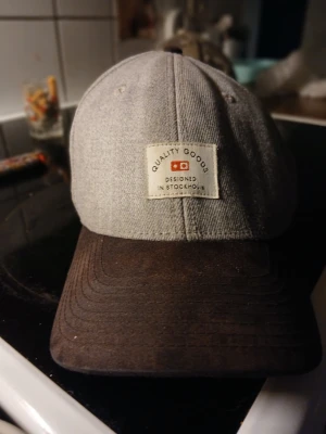 Grå keps med brun skärm - Snygg keps i grått tyg med mörkbrun böjd skärm. Framtill sitter en patch med texten 'Quality Goods Designed in Stockholm' och en liten röd detalj. Klassisk modell som passar till det mesta och ger en clean look.