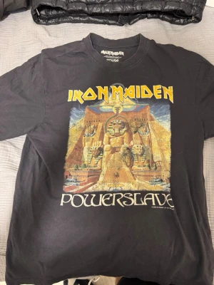 Iron Maiden Powerslave t-shirt svart - Svart t-shirt med Iron Maiden Powerslave-tryck framtill. Klassisk passform med rund hals och korta ärmar. Färgglatt motiv med egyptiskt tema och tydlig Iron Maiden-logga. Perfekt för dig som gillar bandmerch och rockstil.