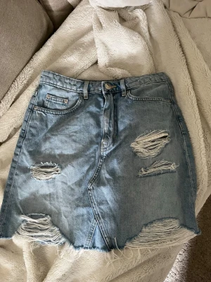 Ljusblå jeanskjol med slitningar - Trendig ljusblå jeanskjol i kort modell med råa kanter och flera slitningar framtill. Kjolen har klassiska fickor både fram och bak samt knapp och dragkedja i midjan. Perfekt för en avslappnad och cool stil.