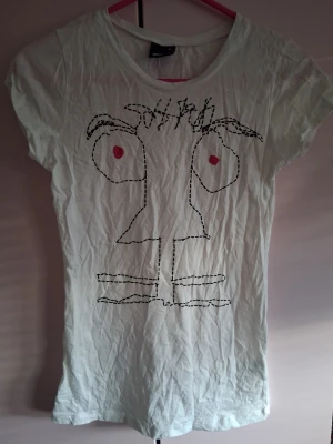 Vit t-shirt med ansiktsmotiv - Vit t-shirt med ett stort, handritat ansiktsmotiv i svart och röda detaljer på framsidan. T-shirten har rund hals och korta ärmar. Tillverkad i mjukt material som känns skönt mot huden. Perfekt för dig som gillar unika prints.