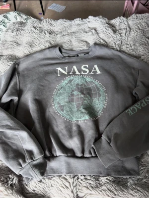 Grå NASA sweatshirt med tryck - Cool grå sweatshirt med stort NASA-tryck och jordglob på bröstet. Tröjan har ribbade muddar vid ärmslut och midja samt texten 'SPACE' på ena ärmen. Perfekt för dig som gillar rymden och streetstyle.