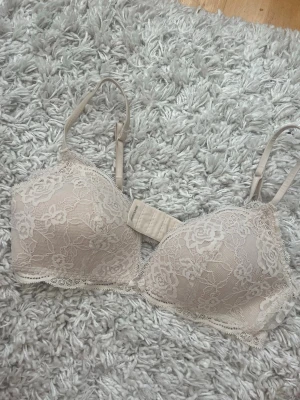 Intimissmi Beige spets-bh med blommönster - Säljer en beige bh med vacker spets och blommönster. Bh:n har justerbara axelband och vadderade kupor för extra komfort. Spetsdetaljerna ger en romantisk känsla och bh:n stängs med hysk- och hakknäppning bak.