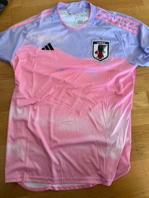 Japan Adidas rosa fotbollströja XL - Säljer en snygg Japan fotbollströja från Adidas i slim fit. Tröjan har en cool färgövergång från ljuslila till rosa, med svarta och rosa detaljer samt Japan-emblem på bröstet. Perfekt för dig som gillar unika matchtröjor och sportig stil.