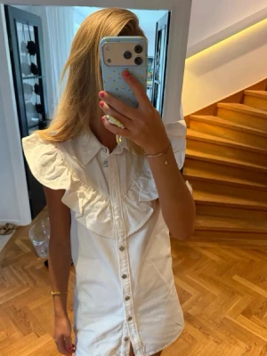 Vit kortklänning jeans med volangdetaljer - Kort vit klänning med knappar framtill och stora volangdetaljer över axlarna. Klänningen har skjortkrage och är ärmlös, vilket ger en fräsch och trendig look. Perfekt för varma dagar och enkel att styla med accessoarer.