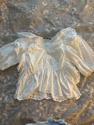 Vit broderad blus från Zara - Söt vit blus från Zara med broderade hål och volanger. Blusen har v-ringning, puffiga trekvartsärmar och en luftig passform. Perfekt till jeans eller kjol för en trendig look.
