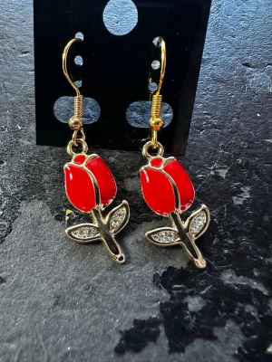 Röda rosörhängen med gulddetaljer - Unika örhängen formade som röda rosor med guldfärgade detaljer och gnistrande stenar i bladen. Hängande modell med krok, perfekta för dig som vill sticka ut med en färgklick och snygg design. Materialet är metall med emalj och små glittrande stenar.