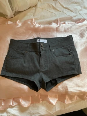 Kostymshorts - Snygga mörkgrå shorts med midwaist passform. Klassisk femficksdesign med två knappar fram och bakfickor med lock och knapp. Perfekta för sommaren och lätta att matcha med olika toppar.