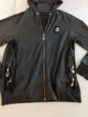 Phillip Plein huvtröja med detaljer - Snygg mörkgrå Philip Plein hoodie med huva och flera silverfärgade dragkedjor på sidorna. På bröstet finns en patch med dödskalle och korslagda ben. Hoodien har coola detaljer och metalliska zippar som ger en edgy vibe. Perfekt för dig som gillar streetstyle.