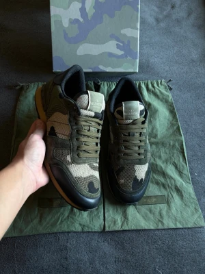 Valentino rockrunners| 42/43 - Snygga sneakers från Valentino med militärgrönt camomönster, svarta detaljer och gummisula. Ovandelen är i mesh och mocka för en cool och sportig look. Perfekta för dig som vill sticka ut med stilrena och trendiga skor.