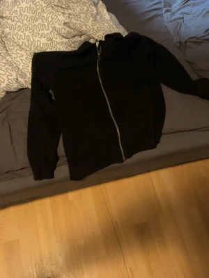 Svart hoodie med dragkedja - En enkel svart hoodie med dragkedja framtill och huva. Hoodien har långa ärmar och ribbade muddar vid ärmslut och nederkant. Perfekt för en avslappnad och stilren look. Materialet ser ut att vara mjukt och bekvämt, troligen bomull eller bomullsblandning.