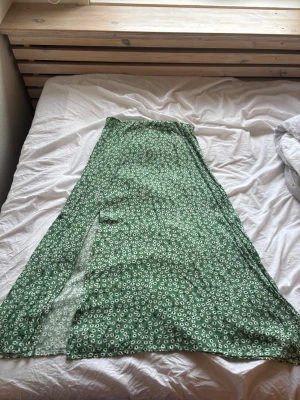 Grön blommig långkjol med slits - Säljer en grön långkjol med vitt blommönster och en snygg slits på sidan. Kjolen har en utsvängd passform och är perfekt för varma dagar. Materialet känns lätt och luftigt, vilket gör den bekväm att bära på sommaren. Använd endast 2 gånger. Nypris är 148 kr mitt pris 70 kr,  köparen står för frakten. 