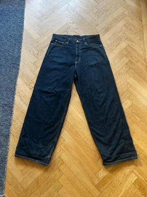 Mörkblåa baggy jeans - Säljer ett par mörkblå jeans med raka, vida ben och hög midja. Klassisk femficksmodell med kontrastsömmar och knappgylf. Jeansen har en enkel och stilren look, perfekta för dig som gillar en avslappnad men trendig stil. Längst ner på benen är de mycket sönder på grund av heelbite. 