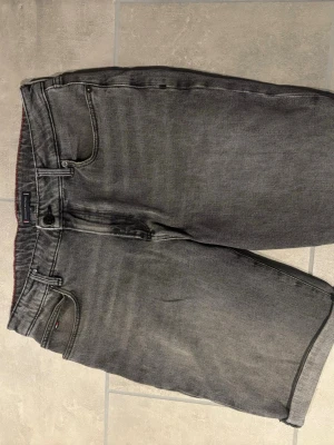 Grå jeansshorts med fem fickor - Säljer ett par grå jeansshorts i klassisk femficksmodell. Shortsen har normal passform, knappgylf och bälteshällor. Tillverkade i bomull med en tvättad look och detaljer som bakfickor och en liten logga på fickan.