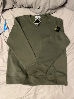 Olivgrön sweatshirt från Stone Island - Snygg olivgrön sweatshirt från Stone Island med klassisk rund hals och ribbade muddar vid ärmslut och nederkant. Tröjan har den ikoniska Stone Island-loggan på vänster ärm och är tillverkad i mjukt bomullsmaterial. Perfekt för en avslappnad streetwear-look.