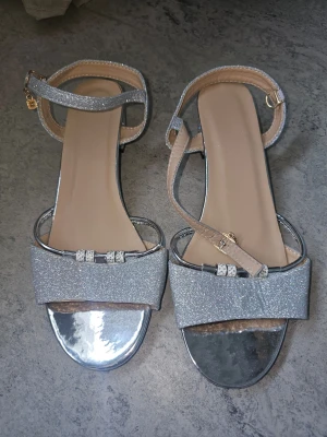 Glittriga silvriga sandaler med klack - Snygga sandaler i silver med glittrig finish och låg klack. Remmarna har spänne runt vristen och dekoration framtill med små stenar. Insidan är beige och sulan har metallic-look. Perfekta för att sticka ut och ge extra bling till outfiten.