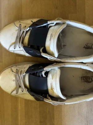 Valentino open  - Snygga vita sneakers från valentino med bred svart rem över vristen och klassisk snörning. Skorna har en låg profil och är tillverkade i läder med gummisula. Perfekta för en stilren och modern look.