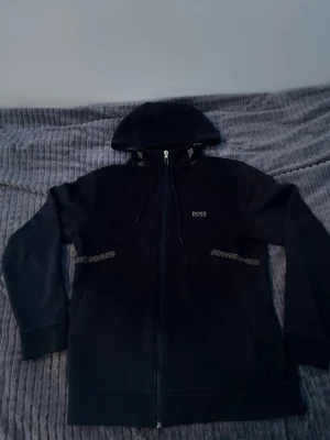 Svart hoodie med dragkedja från BOSS - Snygg svart hoodie från BOSS med dragkedja framtill och huva med snören. Diskret BOSS-logga på bröstet och dekorativa detaljer längs sidorna. Tillverkad i mjukt material som känns skönt mot huden. Perfekt för en avslappnad och stilren look.