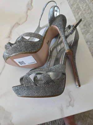 Glittriga silverfärgade platåpumps - Säljer ett par glittriga silverfärgade platåpumps med öppen tå och hög smal klack. Skorna har justerbar rem runt hälen och är tillverkade i syntetmaterial. Perfekta för dig som vill sticka ut och glänsa lite extra.