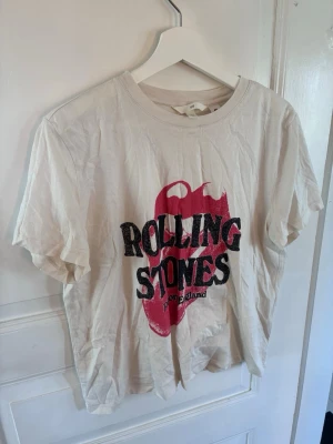Rolling Stones t-shirt från H&M - Vit t-shirt från H&M med Rolling Stones-tryck framtill i svart och rött. Klassisk rund halsringning och korta ärmar. Perfekt för dig som gillar bandtröjor och vill ha en avslappnad stil.