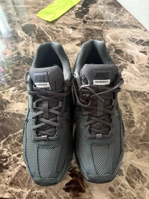Nike Vomero 5 grå sneakers - Nike Vomero 5 sneakers i helgrå färg med mesh och syntetpaneler för extra ventilation och stil. Skorna har en chunky sula och reflekterande detaljer på hälen. Klassisk Nike Swoosh på sidan och snörning framtill. Perfekta för dig som gillar sportig och clean look.