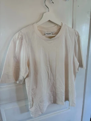 Beige t-shirt från Kappahl - En enkel och stilren beige t-shirt från Kappahl i mjuk bomull. T-shirten har rund halsringning och korta ärmar med lätt puff vid axlarna för en trendig touch. Perfekt basplagg att matcha till olika outfits.