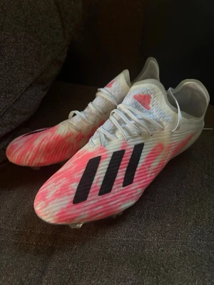 Adidas fotbollsskor - Säljer ett par snygga Adidas fotbollsskor i vitt med rosa detaljer och svarta klassiska stripes. Skorna har snörning och är tillverkade i ett lätt syntetmaterial som ger bra bollkontroll och passform. Perfekta för dig som vill sticka ut på planen!