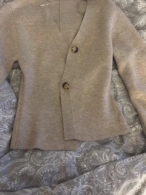 Beige kofta med knappar - Stilren beige kofta med v-ringning och två stora, marmorerade knappar framtill. Koftan har långa ärmar med uppvik och knappdetalj vid ärmslutet. Materialet ser ut att vara mjukt och stickat, perfekt för lager på lager.