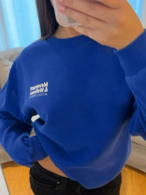 Blå sweatshirt med tryck - Cool blå sweatshirt med vit text framtill och stort tryck på ryggen. Texten handlar om 'Movement & Wellness' och 'Active Lifestyle'. Tröjan har rund hals och ribbade muddar vid ärmar och midja. Perfekt för en avslappnad och sportig look.