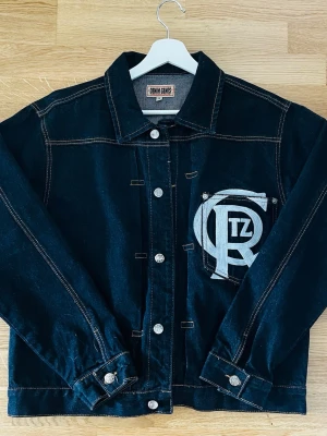 Svart jeansjacka med stor patch - Snygg svart jeansjacka från Cortez med kontrastsömmar och en stor vit patch. Klassisk krage, ganska ny kanpt använd 