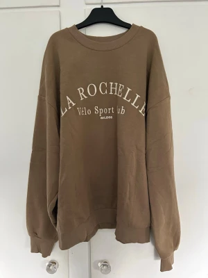 Beige sweatshirt - Säljer en brun/ beige sweatshirt med trycket 'LA ROCHELLE Vélo Sport Club' framtill. Tröjan har rund halsringning, ribbade muddar vid ärmslut och nederkant. Köpt för två år sedan, men endast använt en gång. Nypris: 499kr. 