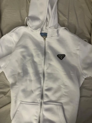Vit hoodie med dragkedja från Prada - Säljer en clean vit hoodie från Prada med dragkedja framtill och klassisk huva. Hoodien har en ficka på magen och den ikoniska svarta Prada-loggan i metall på bröstet. Perfekt för dig som gillar exklusiv streetwear och vill sticka ut med en lyxig touch.