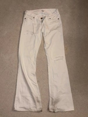 Vita bootcut jeans med slitningar - Säljer ett par vita bootcut jeans med slitna detaljer vid knäna och femficksmodell. Jeansen har låg midja och vida ben, perfekta för en avslappnad och trendig look. Stängs med knapp och dragkedja framtill. KOM PRIVAT FÖR ATT SE BYXORNA LÄNGST NER, DE HAR VARIT LÅNGA PÅ MIG SÅ DE HAR SLÄPATS MEN KAN ENKELT KLIPPA BORT! 