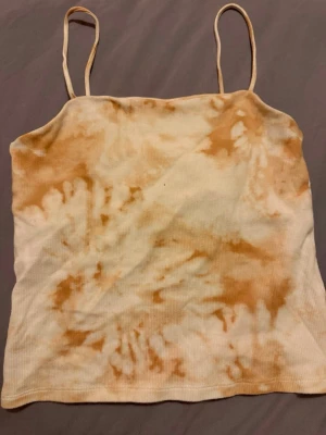 Beige tie-dye ribbad linne - Säljer ett ribbat linne med tunna axelband i beige och vitt tie-dye mönster. Toppen har en rak skärning och är stretchig, perfekt för en avslappnad och trendig look.