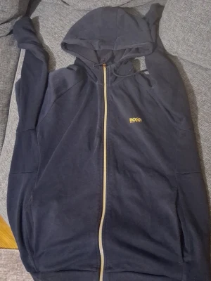 Mörkblå hoodie med dragkedja från BOSS - Snygg mörkblå hoodie från BOSS med gul logga på bröstet och ryggen. Hoodien har dragkedja framtill, huva med dragsko och två sidofickor. Tillverkad i mjukt material som känns skönt mot huden. Perfekt för en avslappnad och stilren look.