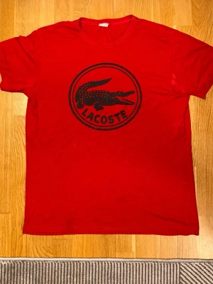Röd Lacoste t-shirt med tryck - Röd t-shirt från Lacoste med stort svart krokodiltryck och logga på bröstet. Klassisk rund hals och korta ärmar. Tillverkad i mjuk bomull för skön känsla och enkel stil.