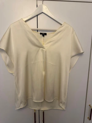 Krämvit blus från C&A - Snygg krämvit blus från C&A med v-ringning och korta ärmar. Tillverkad i ett glansigt, mjukt material som ger en lyxig känsla. Perfekt att styla med jeans eller kjol för en trendig look.