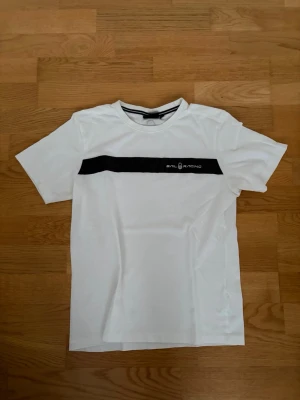 Vit Sail Racing t-shirt med svart tryck - Fräsch vit t-shirt från Sail Racing med en svart horisontell rand och logga över bröstet. Klassisk rund hals och korta ärmar. Tillverkad i mjuk bomull som känns skön mot huden. Perfekt till jeans eller shorts för en clean look. Storleken motsvarar S/XS i herr. Pris kan alltid diskuteras! Tveka inte på att höra av dig vid frågor eller funderingar kring storlek eller annat. Använd endast ett fåtal gånger!