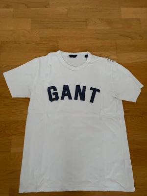 Vit GANT t-shirt med blå broderad logga - Vit t-shirt från GANT med stor blå logotyp i text på bröstet. Klassisk passform med rund halsringning och korta ärmar. Tillverkad i mjuk bomull som känns skön mot huden. Perfekt för en clean och enkel stil. Storleken passar herr S.