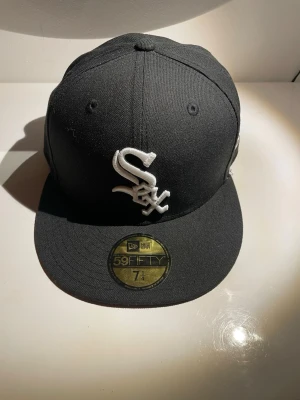 Svart New Era 59FIFTY White Sox keps, Size 7 1/4 - Snygg svart keps från New Era 59FIFTY med Chicago White Sox-logga broderad i vitt framtill. Klassisk flat peak och broderad New Era-logga på sidan. Tillverkad i ett robust tyg som håller formen. Perfekt för dig som gillar streetwear och sportig stil.
