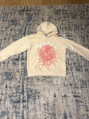 Beige hoodie med spindelnätsprint - Snygg beige hoodie med huva och ett stort rött spindelnätsmönster framtill samt texten 'spider' i vitt ovanför. Hoodien har klassisk passform och ribbade muddar vid ärmslut och nederkant. Perfekt för dig som gillar streetwear och coola tryck.