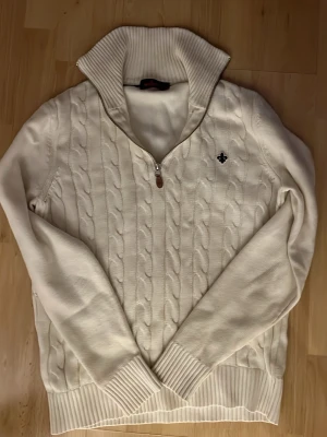 Morris Halfzip vit - Kabelstickad vit tröja från Morris med halv dragkedja och hög krage. Tröjan har ribbade muddar och nederkant samt en liten broderad logga på bröstet. Perfekt för dig som gillar klassisk och stilren design.