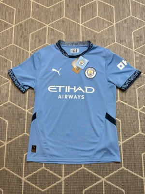 Manchester City Puma - Snygg ljusblå Manchester City matchtröja från Puma med klubbmärke på bröstet och Etihad Airways-tryck framtill. Kort ärm med mörkblå detaljer och text på ärmslut. Tillverkad i lätt och funktionellt material, perfekt för fotboll eller träning.
