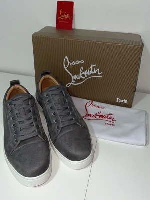 Gråa sneakers från Christian Louboutin - Snygga gråa sneakers från Christian Louboutin med klassisk röd sula och vita kanter. Skorna har ovandel i mocka, matchande gråa skosnören och diskreta sömdetaljer. Perfekta för dig som vill ha en exklusiv och trendig look.