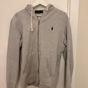 Grå hoodie från Polo Ralph Lauren - Snygg grå hoodie från Polo Ralph Lauren med dragkedja och huva. Tröjan har snörning i huvan, två fickor fram och den klassiska Polo-loggan broderad på bröstet. Perfekt för chill dagar och enkel att matcha med jeans eller joggers.