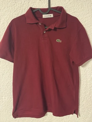 Vinröd pikétröja från Lacoste - Klassisk vinröd pikétröja från Lacoste med korta ärmar och krage. Tröjan har två knappar framtill och den ikoniska gröna krokodilloggan på bröstet och höger arm. Tillverkad i bomull för en bekväm och luftig känsla. Storlek M passar även S, Kan gå ner i pris vid snabb affär!