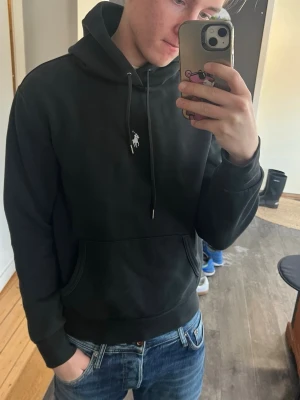 Svart hoodie från Ralph Lauren - Snygg svart hoodie från Ralph Lauren med klassisk vit logga på bröstet. Hoodien har huva med dragsko och en stor magficka. Perfekt för en chill och stilren look. Bara nåt väldigt lite sliten vid fickorna